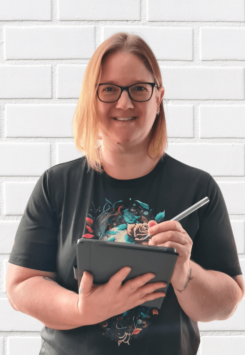 A blonde woman standing in front of a white wall holding a tablet, smiling at the camera